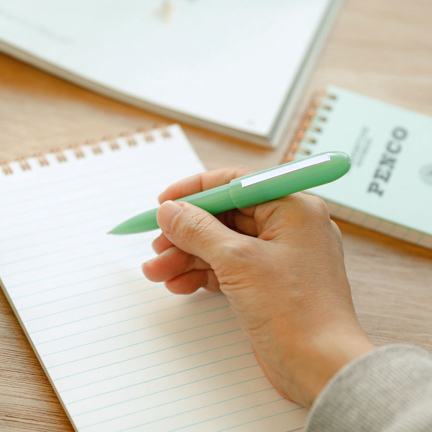right hand with a mint bullet pen ready to write in a lined notebook, other paper and Penco notebooks in the table